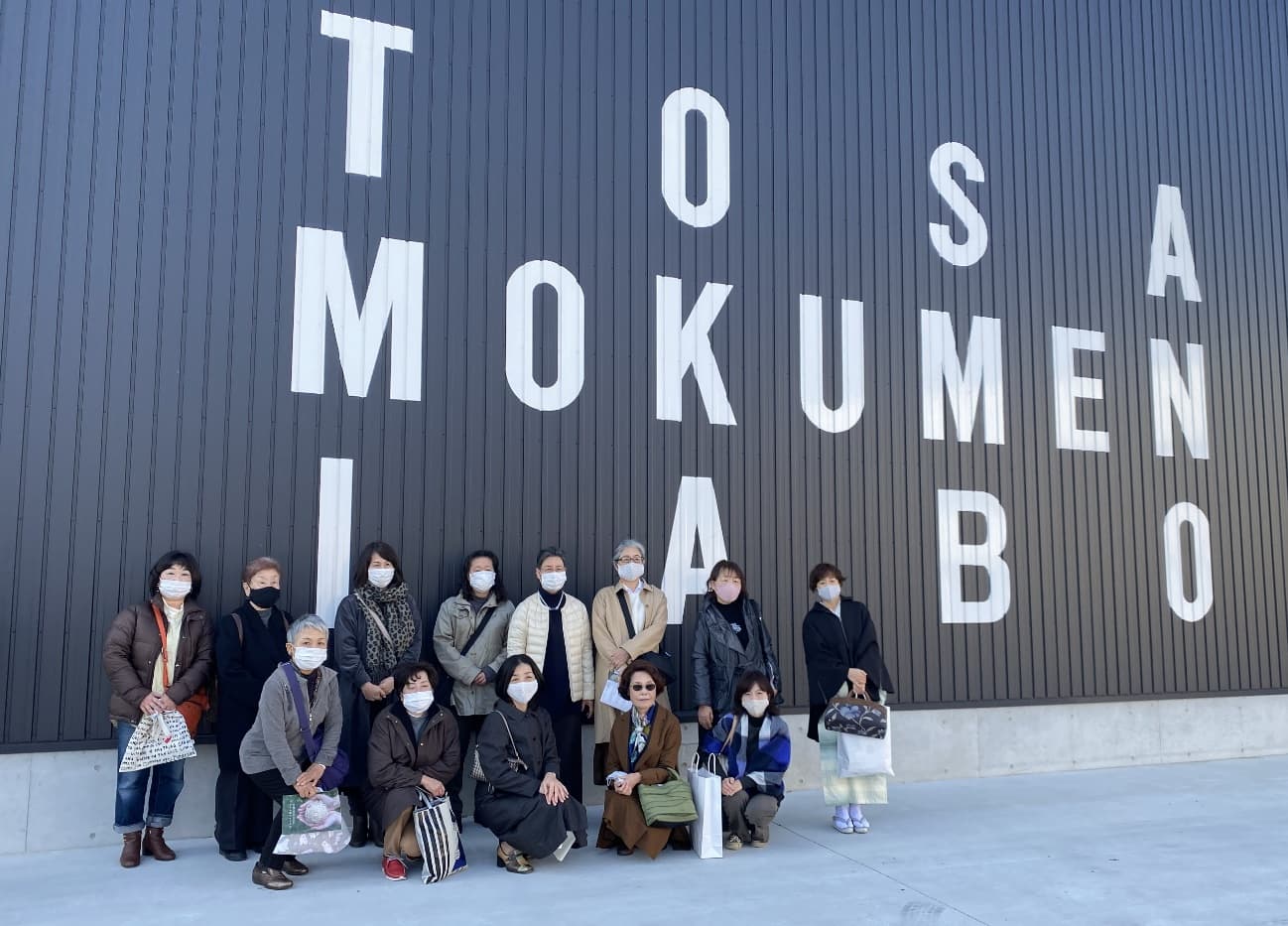 戸田商行の工場前で見学参加者が集合し、建物ロゴを背景に記念撮影する様子